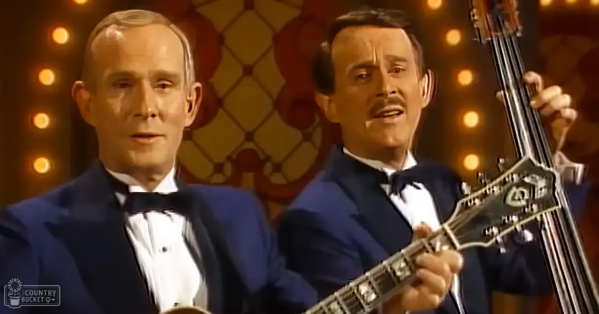 Musical performance scene with two adult male musicians wearing dark blue tuxedos, holding a large stringed instrument against an ornate backdrop with warm-toned lights, set indoors at night.