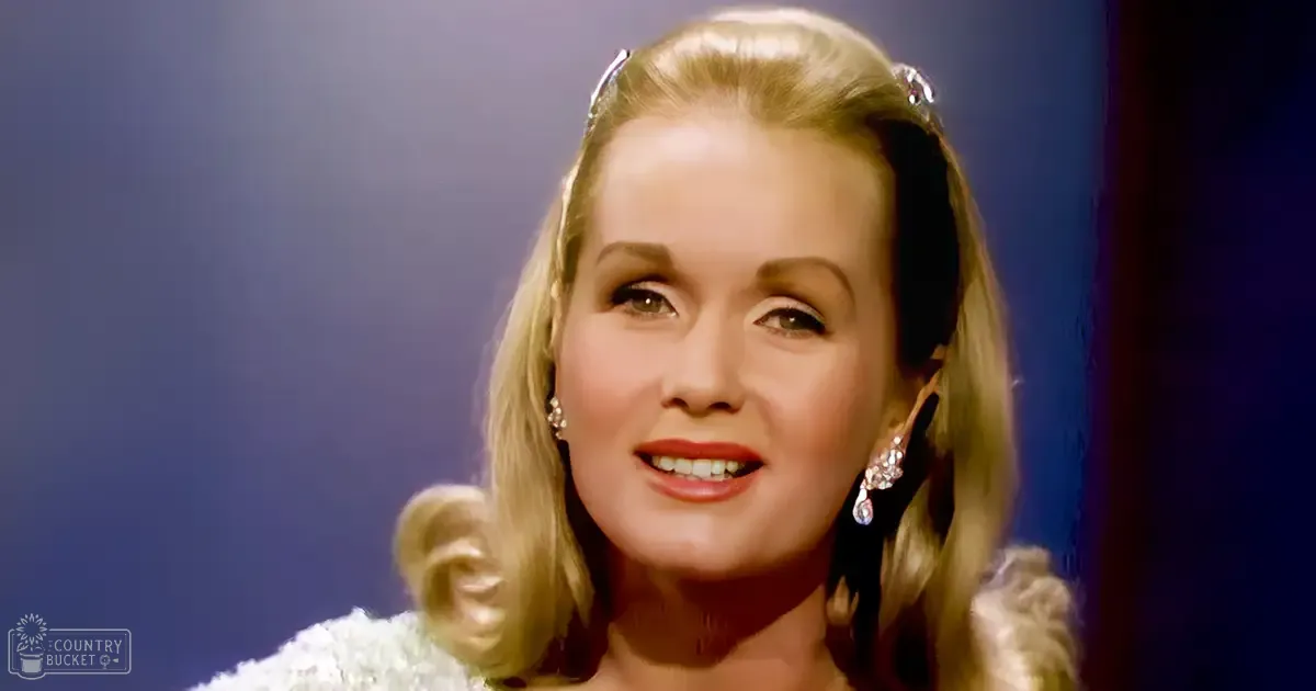 A middle-aged woman in a formal white dress with intricate detailing and large dangling earrings indoors under dark blue lighting.