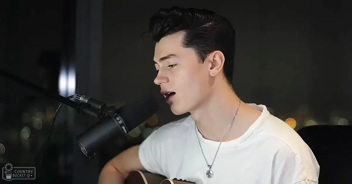 Young adult male in a white t-shirt with short sleeves, standing indoors under dark lighting. Microphone nearby suggests a recording or performance setting.