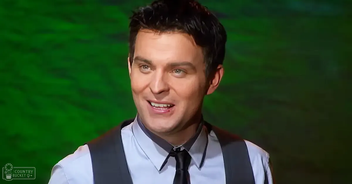 A young adult male in a formal outfit, wearing a light blue dress shirt with a dark collar and black tie, set against a green screen background used for video production.