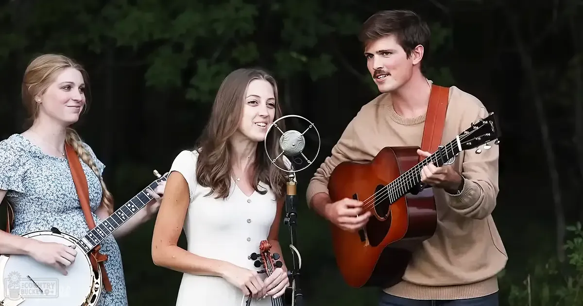 Young adult females and a male performing music outdoors in an evening setting, surrounded by natural greenery.