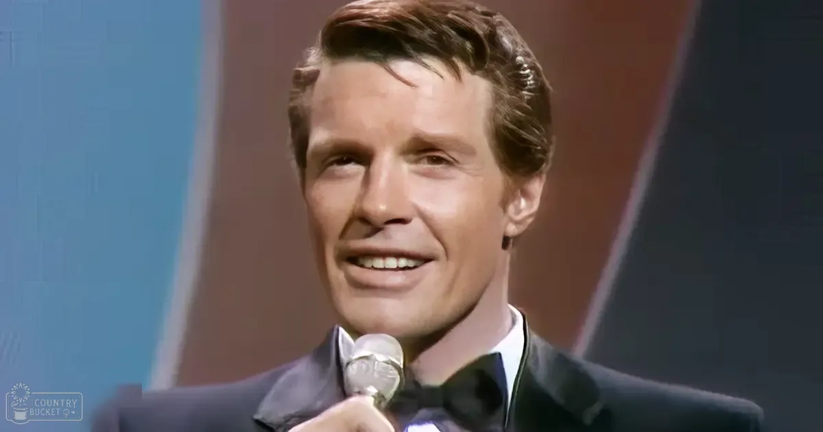 An adult male in a dark blue or black tailored suit from the 1960s, holding a microphone on stage with muted background colors.