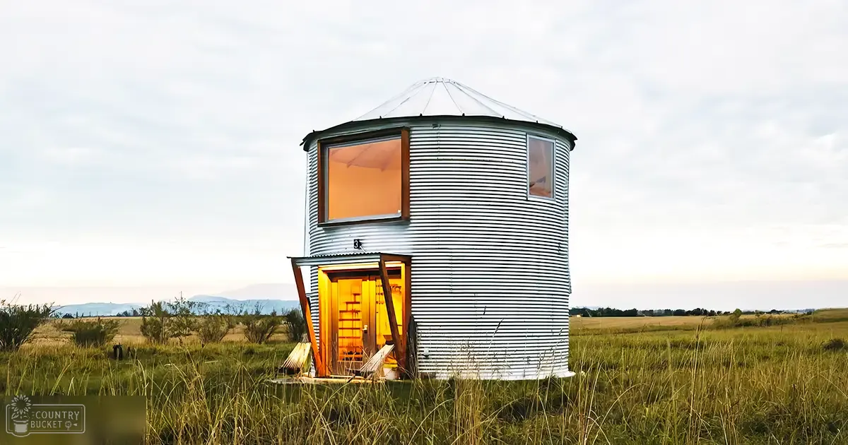 Repurposed grain silo converted into a home in an open field; small cylindrical metal structure with conical roof and large windows, warm lighting visible.