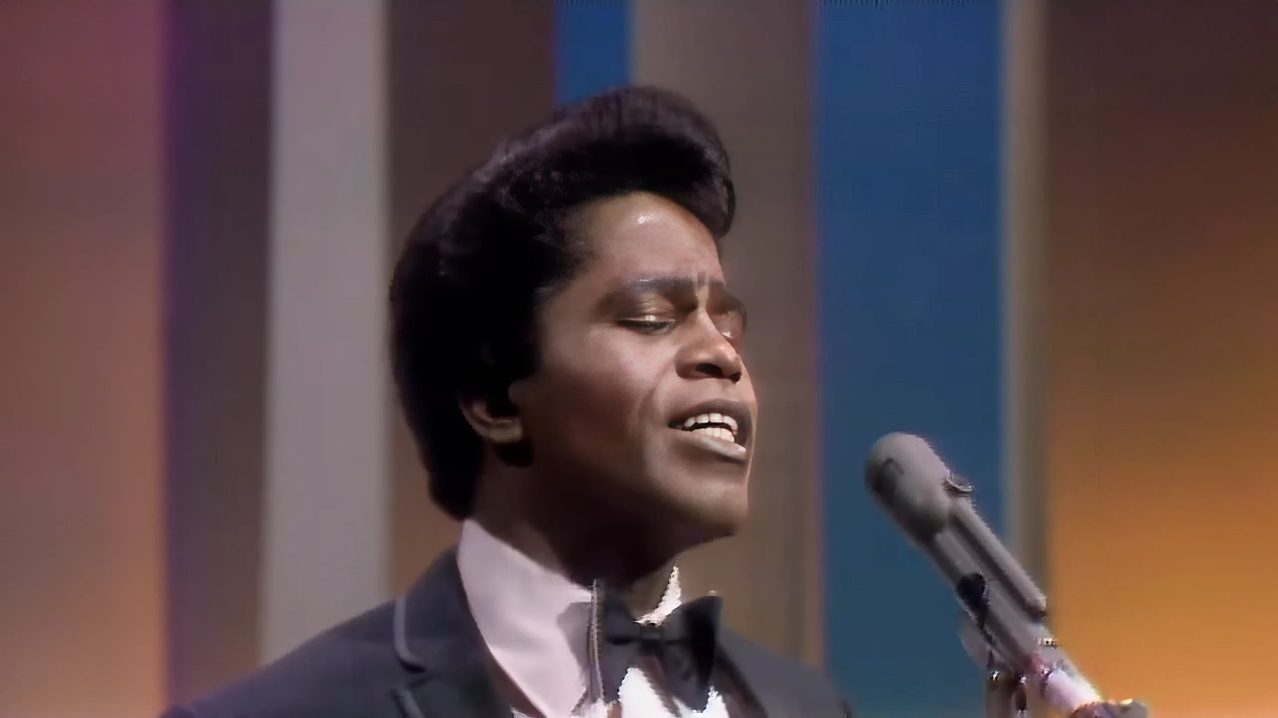 A young adult male singer performing on stage, dressed formally in a dark suit with a white shirt and black bow tie, surrounded by professional stage lighting.
