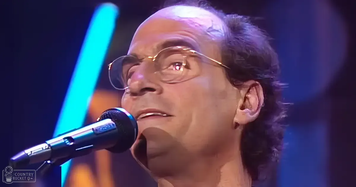 A middle-aged adult male singer wearing a dark-colored shirt, glasses with thin frames, and holding a microphone at what seems to be a concert or performance setting with stage lighting in the background.