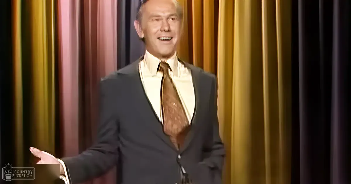 A middle-aged man wearing a dark suit with a white shirt and patterned tie stands on stage, behind a curtain backdrop of vertical gold-brown stripes.