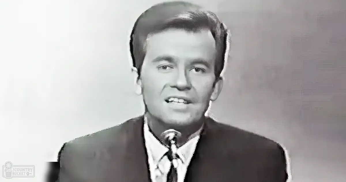 Young adult male singer wearing a dark suit jacket with patterned tie, holding a microphone against his neck, set in an indoor studio environment.