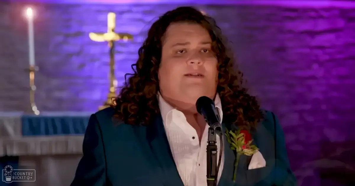 A young adult male in a dark teal suit jacket and white dress shirt stands at a microphone, giving a speech against a stone wall illuminated with purple lighting. A golden cross structure is visible in the background.