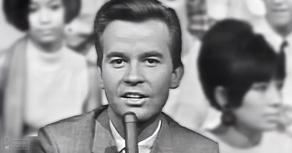 A young adult male in a formal suit jacket and collared shirt stands on an indoor stage with blurred background elements suggesting a mid-20th century performance setting.