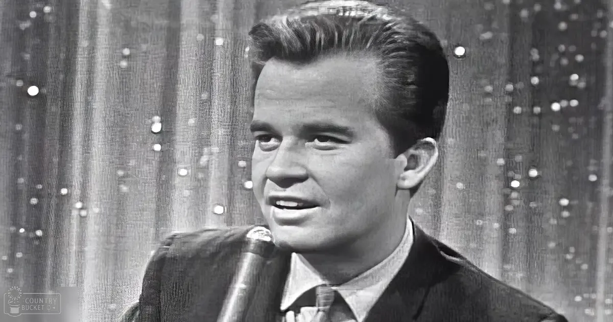 A young adult male in a tailored dark suit and light-colored shirt, standing against an indoor stage backdrop with small evenly spaced lights.