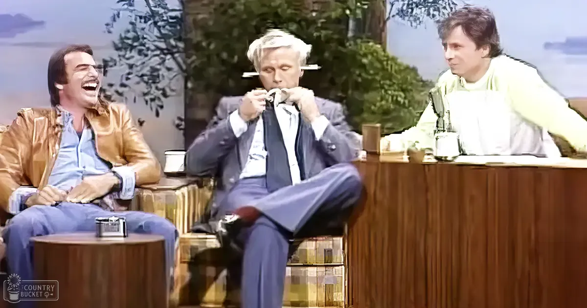 Three individuals in a talk show set from the 1970s, wearing gold leather jackets, dark suits, and green sweaters; studio backdrop of an outdoor scene with trees.