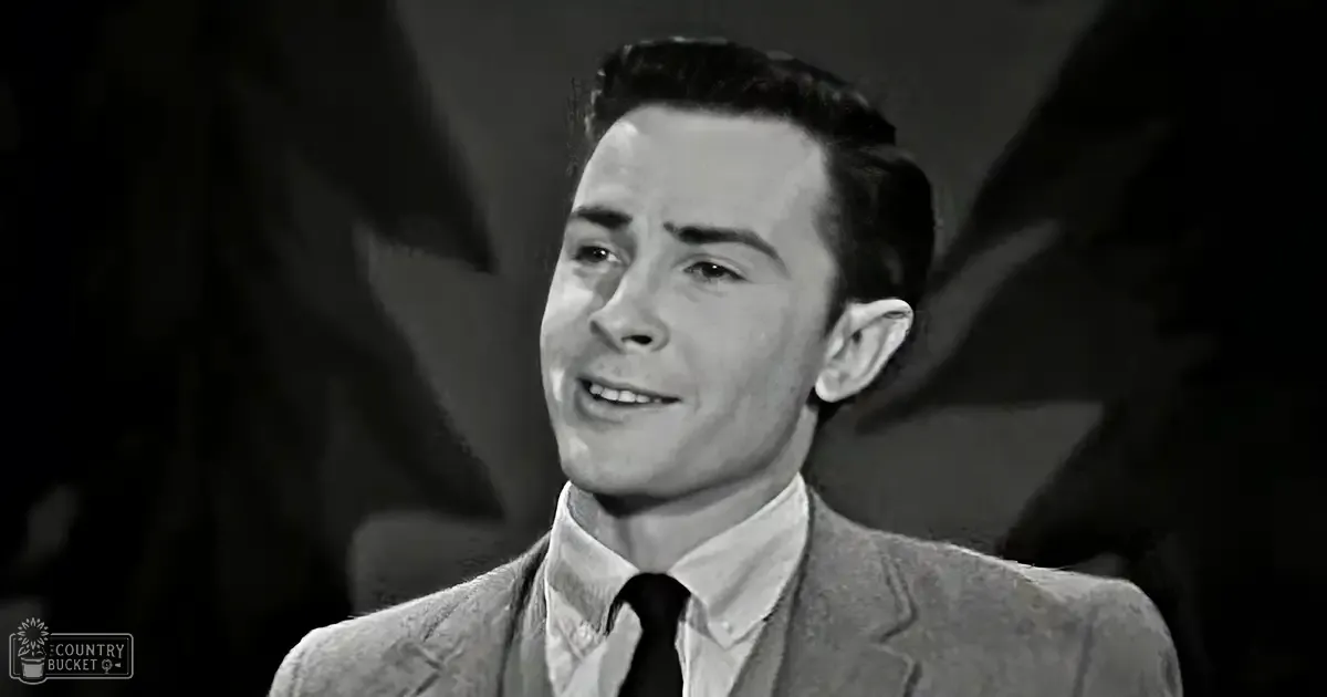 Young adult male wearing a light-colored textured suit jacket with a notched lapel, collared shirt, and dark tie in an indoor setting.