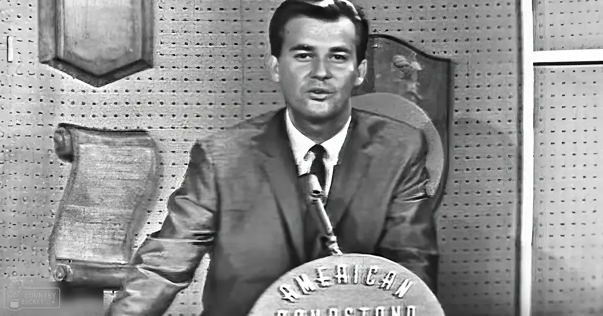 A young adult male in a classic suit jacket and tie, holding a microphone against an indoor studio backdrop with pegboard walls and framed decorative items.