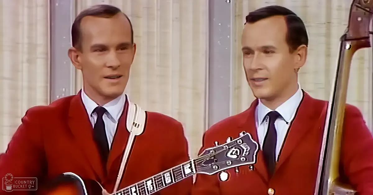 Two adult male singers in red suits perform indoors, holding musical instruments with a bright and even lighting.