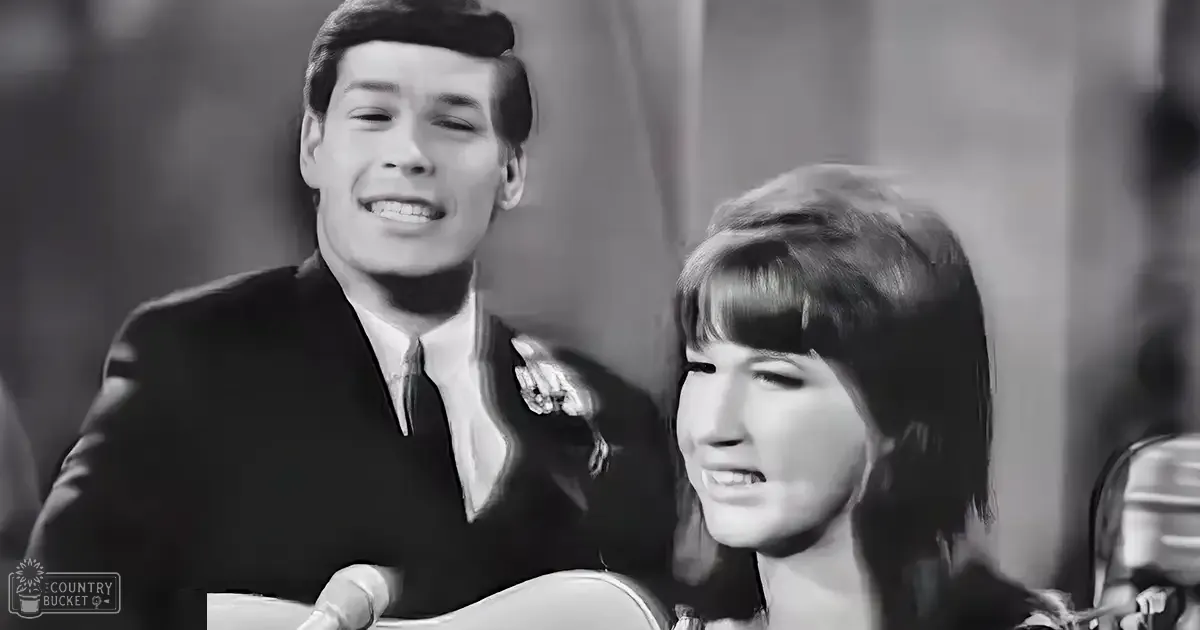 A musical performance scene with two individuals dressed formally against a blurred indoor backdrop suggesting stage or studio lighting.