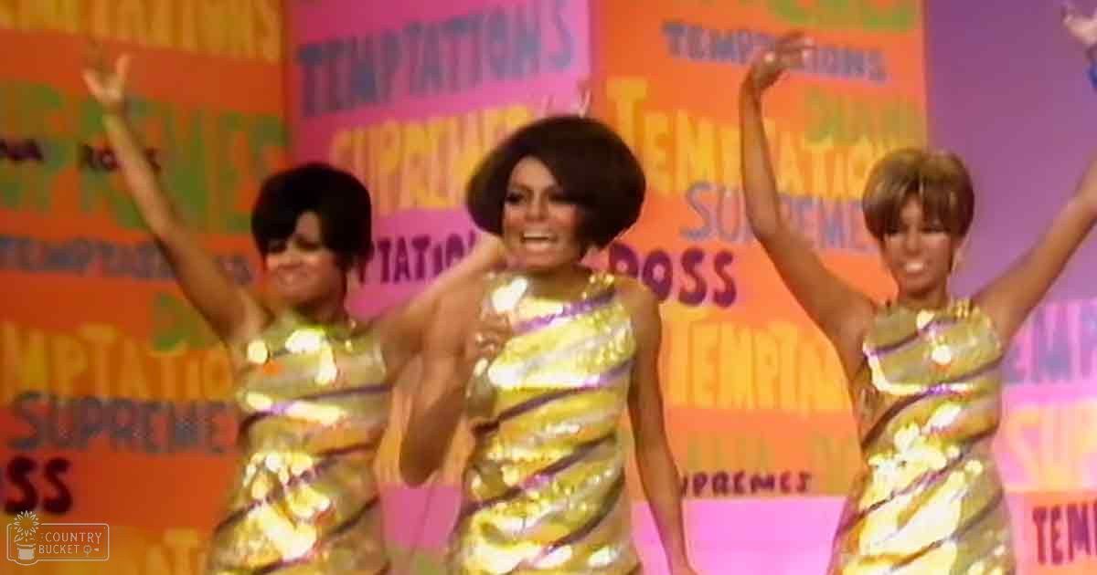 Three adult female performers in shiny, gold-colored dresses with striped patterns on a stage-like setup with 'Temptations' and 'Supremes' backdrop.