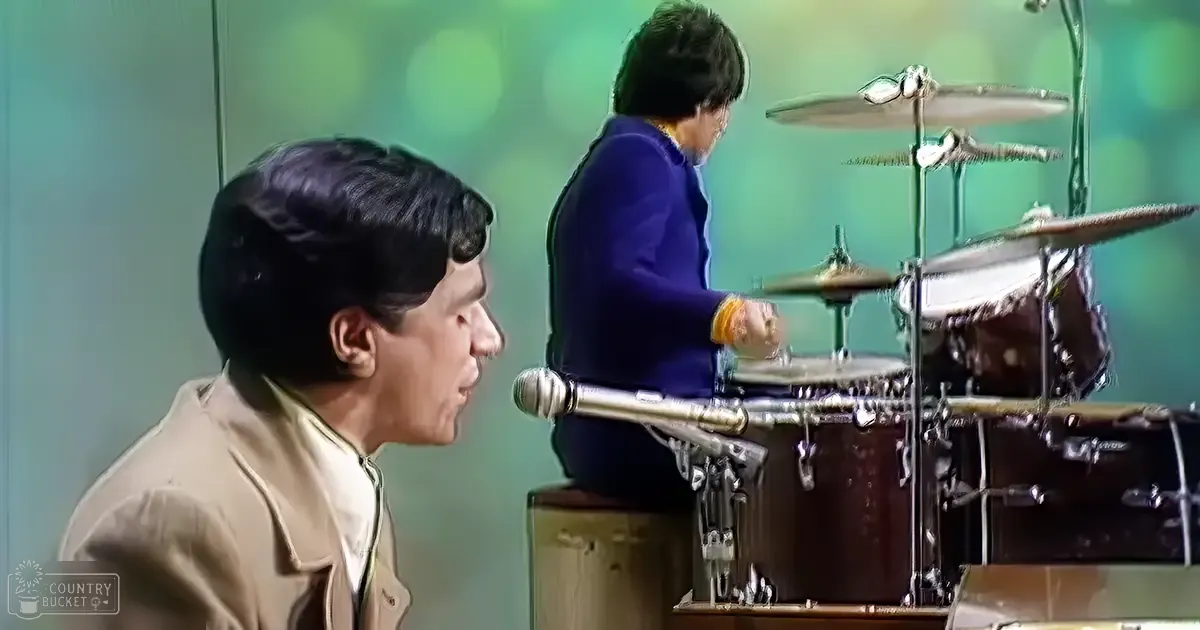 Singer and drummer performing on stage with a drum set in front of a greenish-blue backdrop, likely at an indoor music venue.