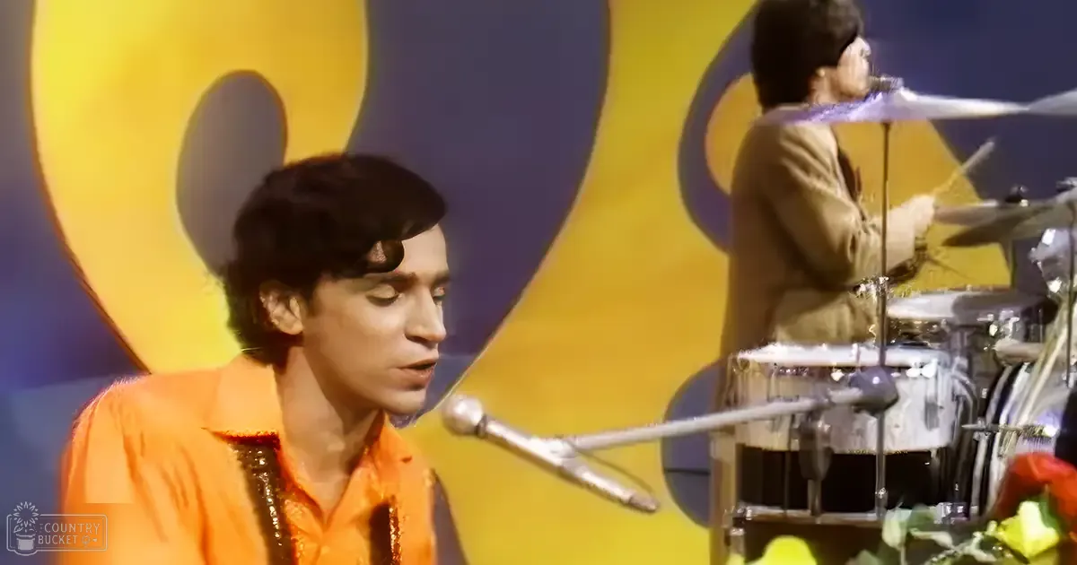 A young adult male singer wearing an orange high-collared shirt with shiny fabric, performing on stage in front of a drum set alongside another young adult male drummer dressed in a light brown jacket and white gloves. The setting is indoors at what appears to be a music event or concert with bright artificial lighting.