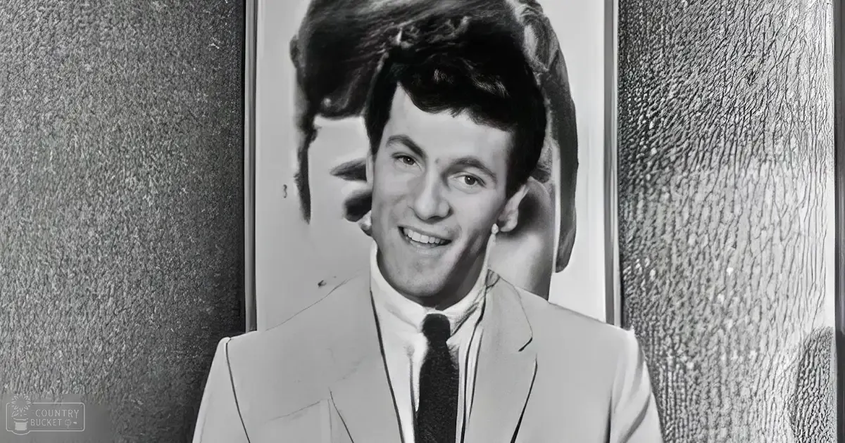 A young adult male in a light-colored suit jacket with a dark tie, wearing a white shirt, stands indoors against textured walls suggesting an office or studio environment.