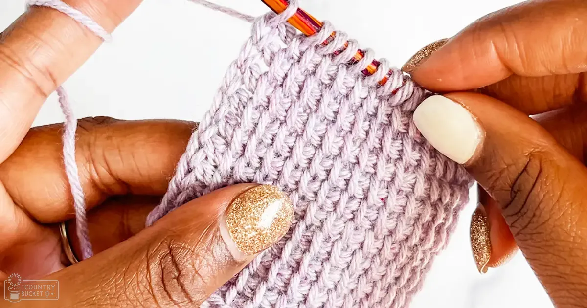 Close-up of hands knitting with yarn, orange-red striped crochet hook, light purple yarn, gold glittery nail polish on one hand, white nail polish and small tool on the other in an indoor setting.