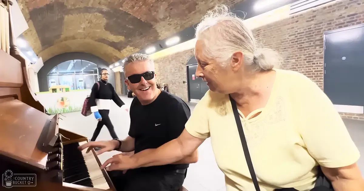 Adult male in black T-shirt playing piano with older adult woman beside him, surrounded by an industrial setting with brick walls and artificial lighting.