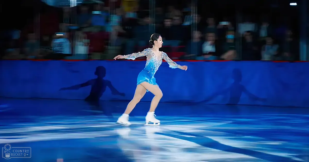 A young adult female figure skater in a blue costume with silver embellishments performs on an indoor ice rink. Blurred spectators are visible in the background, suggesting it's likely during a performance or competition.