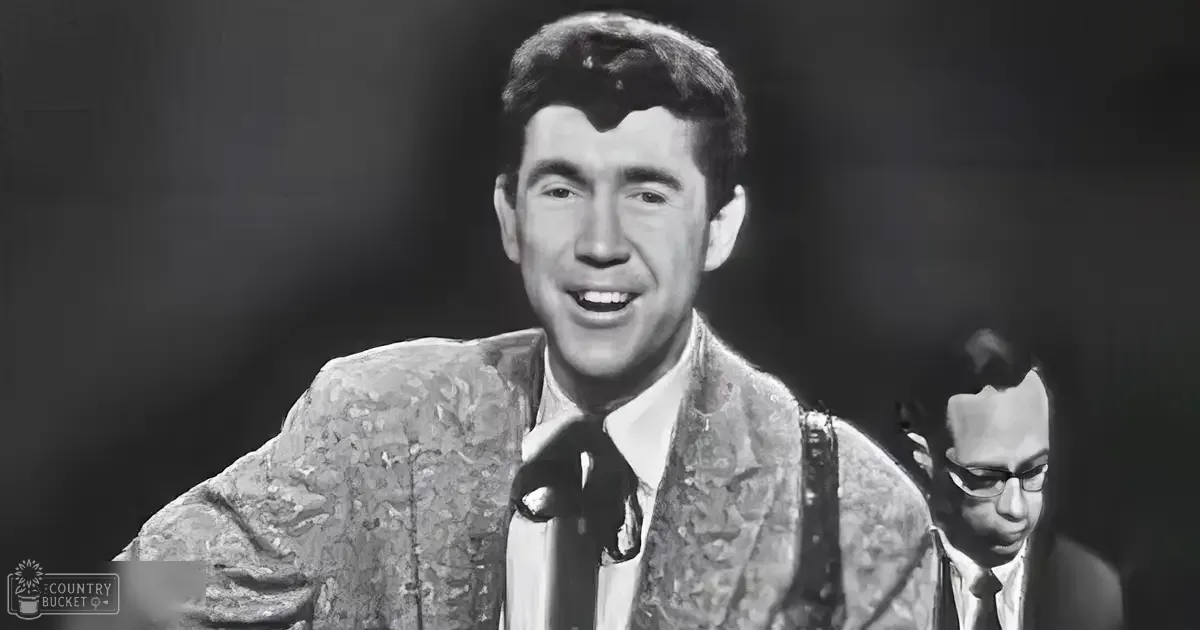 Young adult male in patterned suit jacket on stage with another young adult male in similar attire; dark and out-of-focus background suggests a spotlight effect typical of performances.