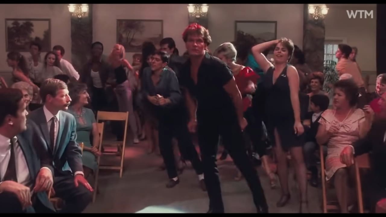 Adult male dancer wearing a black V-neck shirt, leather boots, and red cap, surrounded by spectators in formal attire at an indoor social gathering with warm lighting.