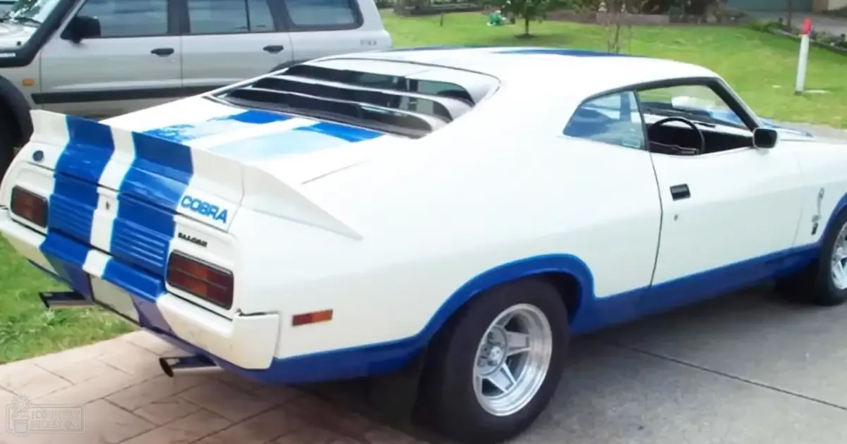Classic Ford Mustang Cobra with white and blue racing stripes, parked on a paved surface amidst a residential area during clear daytime conditions.