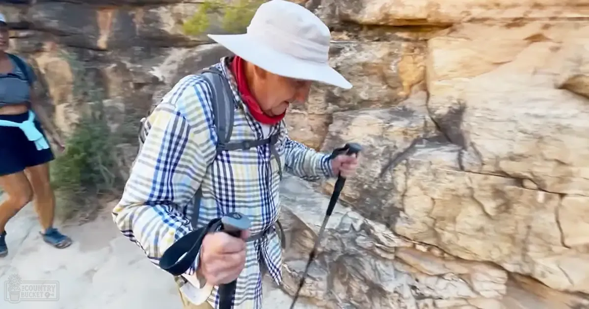 A middle-aged woman wearing a white hat and plaid shirt hikes along a rocky trail, carrying a backpack with trekking poles. A young adult female is visible to the left in athletic attire.