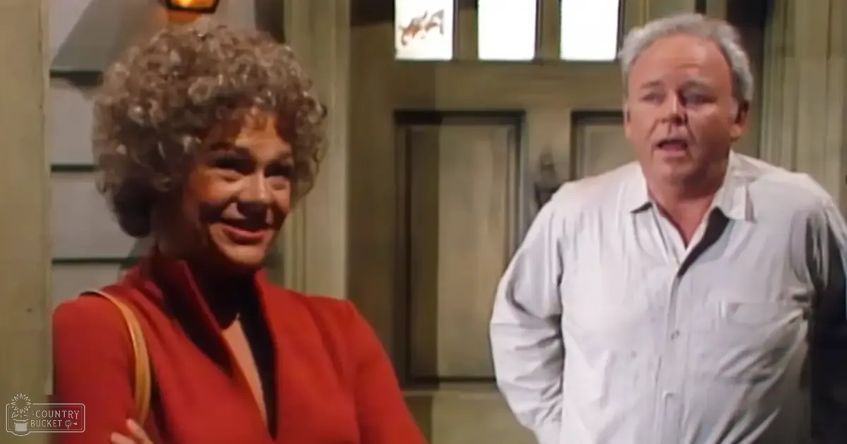 An older adult female in a red sleeveless top with black inner layer, paired with a light brown shoulder bag strap, standing indoors alongside a middle-aged man wearing a white button-up shirt. The setting appears to be an indoor space with columns and doors, possibly a hallway or lobby of a building.