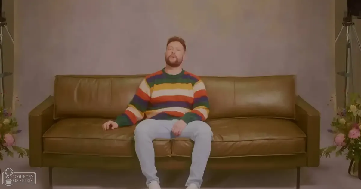 A middle-aged adult male sitting on a brown leather couch in an indoor space with floral decorations, wearing a striped sweater and jeans.