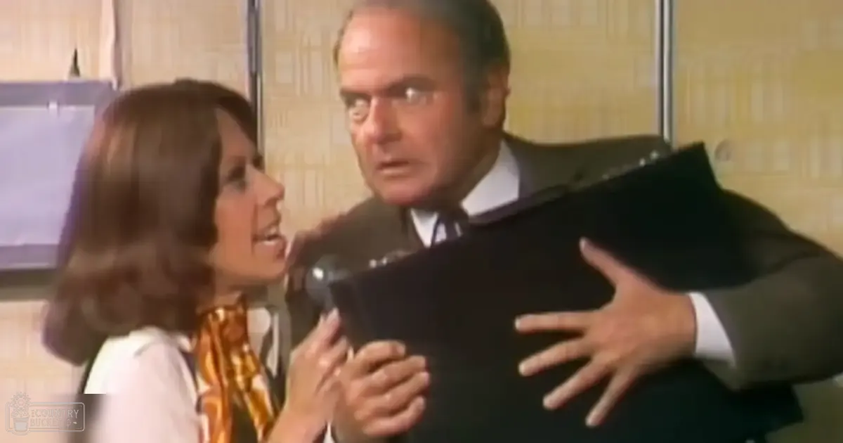 A middle-aged man in a well-fitted dark suit jacket, white shirt, and tie stands in an office setting with a young adult to middle-aged woman wearing a patterned orange and yellow scarf. The scene is set indoors, likely from the mid-20th century.
