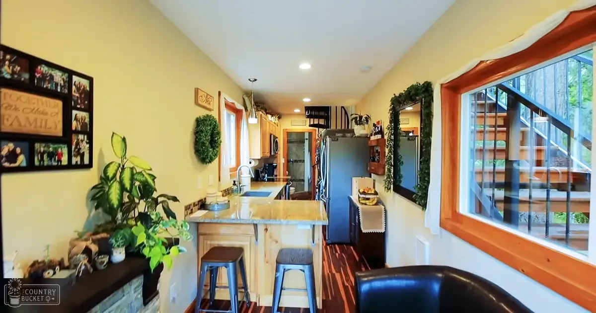 A kitchen area with two bar stools at an island counter, a refrigerator against one wall, and family photos on the left wall above a fireplace mantel adorned with plants.