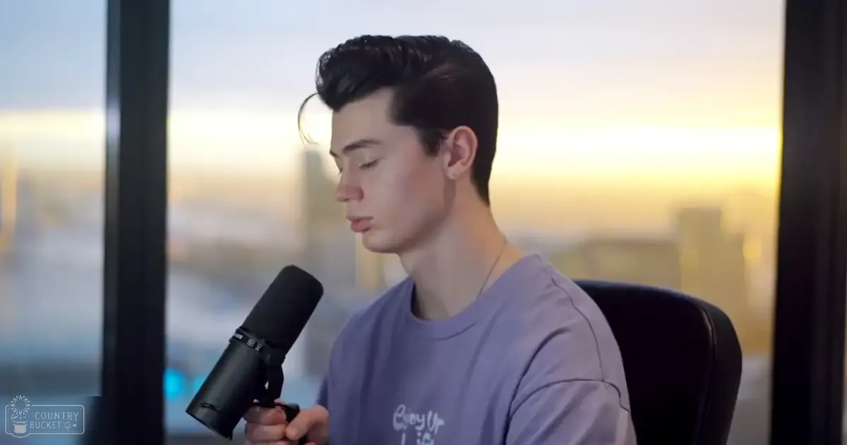 A young adult male wearing a relaxed fit, solid light purple crew neck T-shirt with a cotton blend material, holding a microphone in an indoor setting overlooking a cityscape at sunset or sunrise.