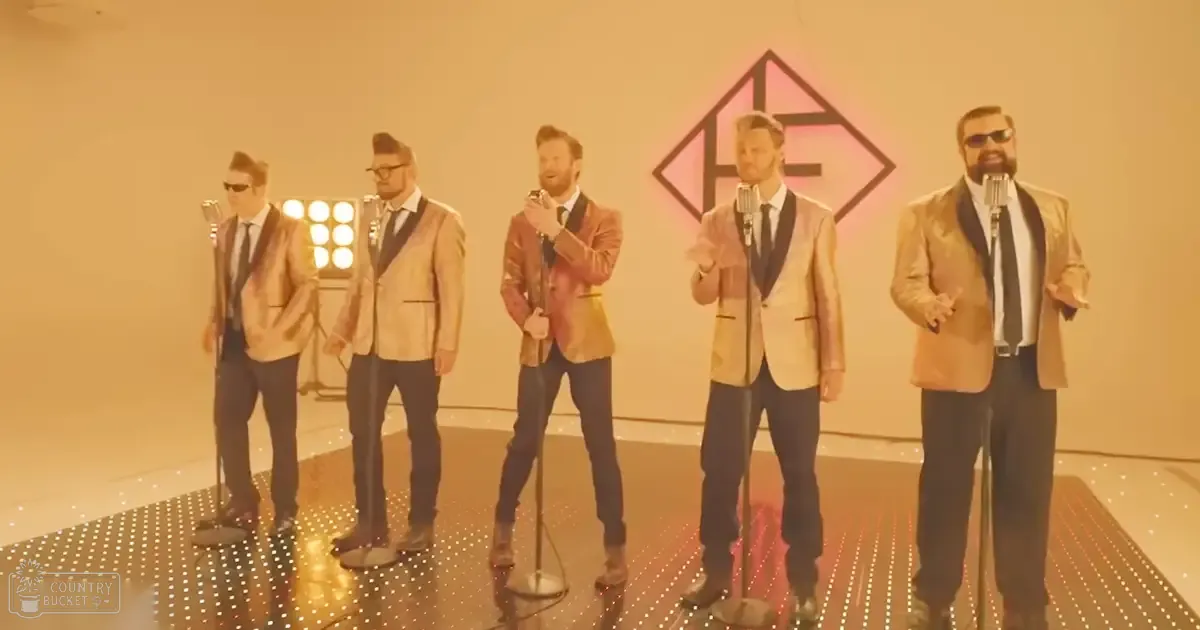 Group of adult male performers in a studio, wearing light brown suit jackets with darker lapels, dark pants, and dark shoes; one holding a microphone.