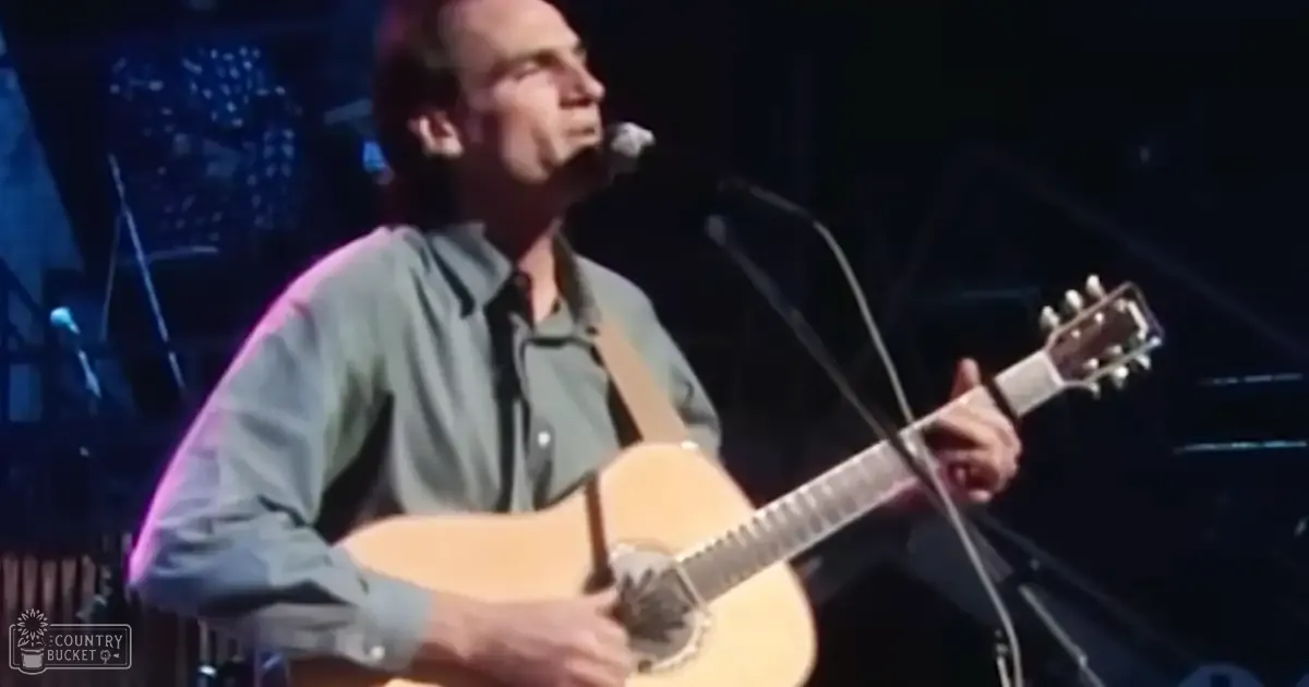 A middle-aged adult male in a dark green, long-sleeved button-up shirt performs on stage with an acoustic guitar under dim indoor lighting.