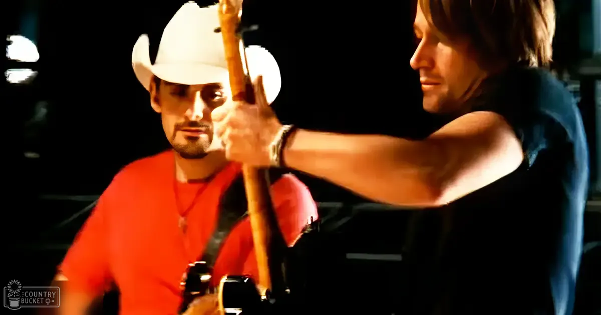 Two adults, one wearing a red t-shirt with a zipper and white cowboy hat, the other in a dark sleeveless shirt with a watch, performing indoors under stage lights.