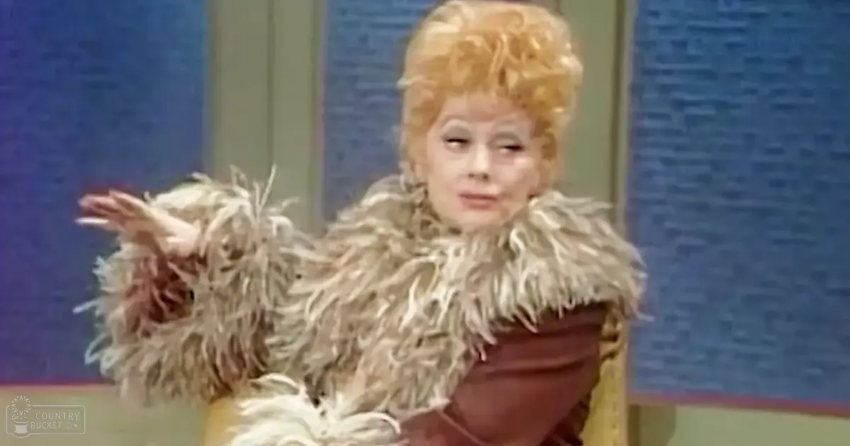An adult female performer in a theatrical setting, wearing a fluffy light brown fur coat with a rich maroon or dark red bodice, indoors on an even-lit stage.