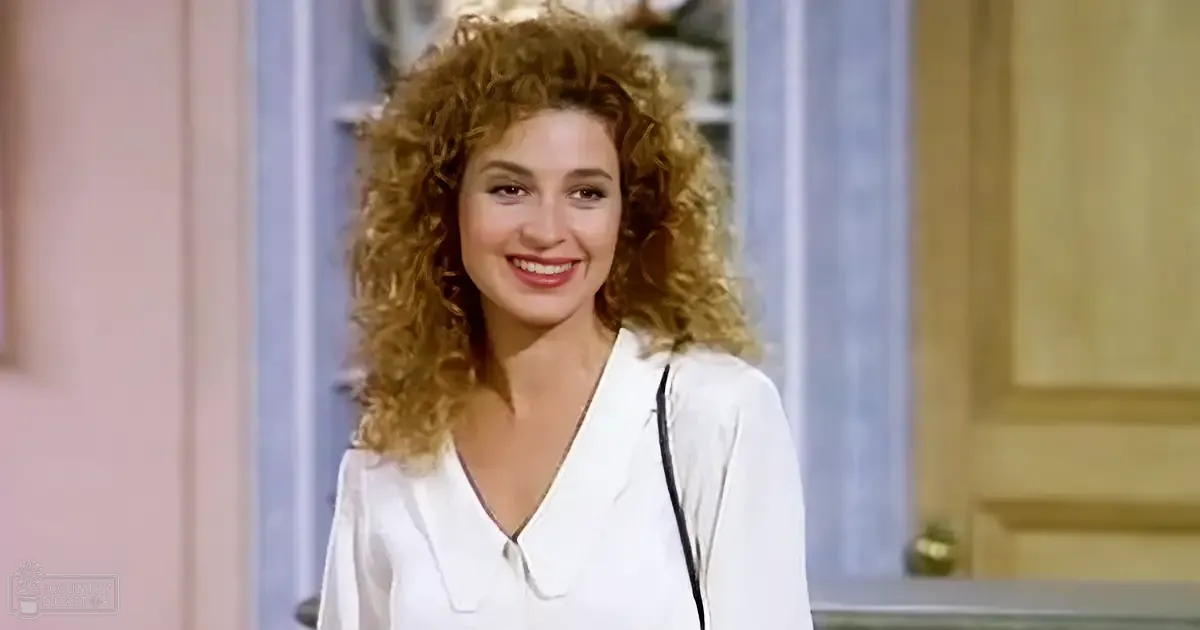 A young adult female in a relaxed, white v-neck blouse with button detail at center of neckline, indoors with light-colored walls and decorative elements.