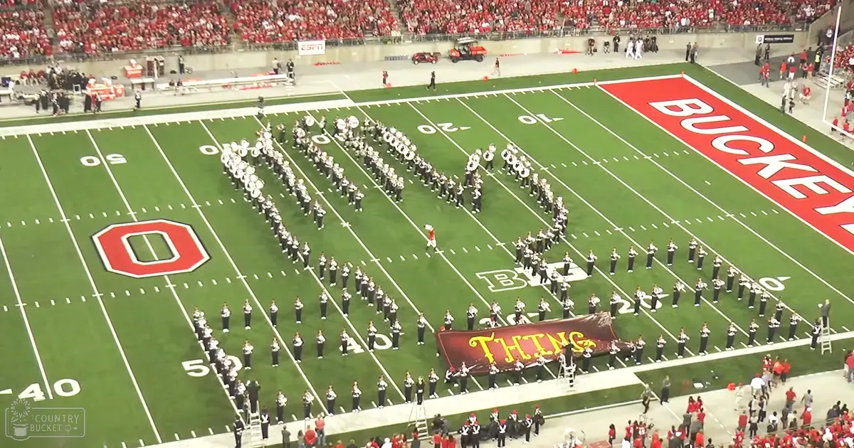 A marching band at Ohio State University's football stadium with young adults in dark uniforms, a red golf cart, and a large 'O' logo of the university.