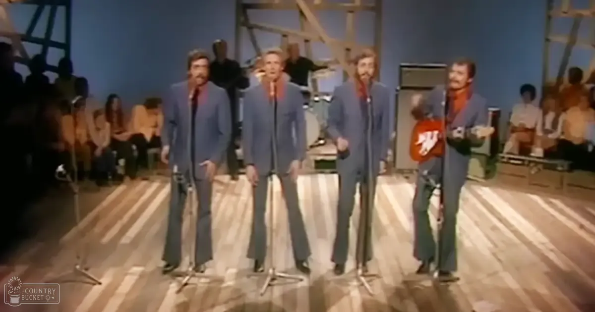 A musical performance group in a light blue suit jacket, red shirt, dark trousers, with an acoustic guitar on stage in front of an audience.