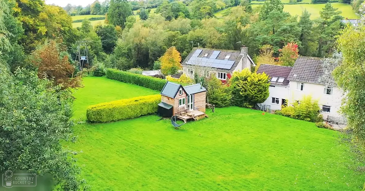 A serene rural landscape with a well-maintained lawn, small wooden structures, and trees with changing foliage in late afternoon light.