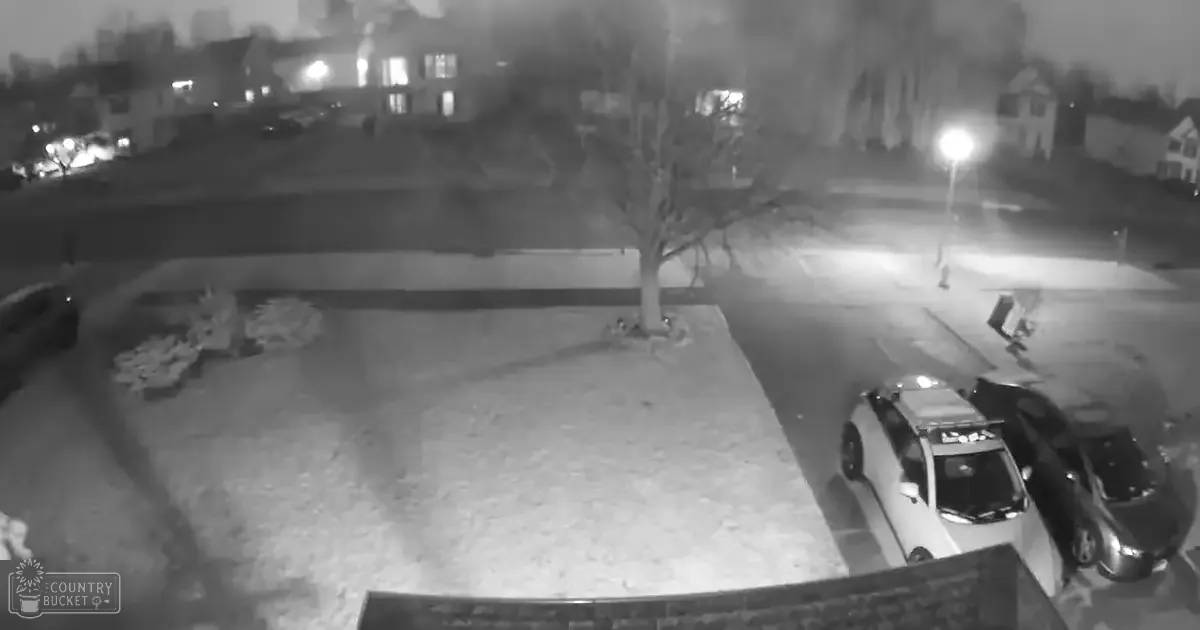 A residential neighborhood at night with illuminated houses, parked cars, and snow-covered yards under artificial light.