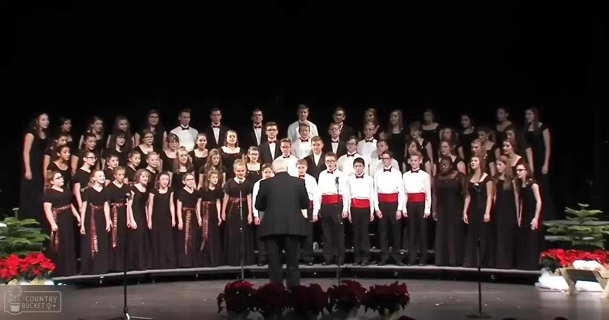 A children's choir performance in a school auditorium, with children wearing black dresses and white shirts paired with black pants, set against a backdrop of dark curtains and stage lighting.