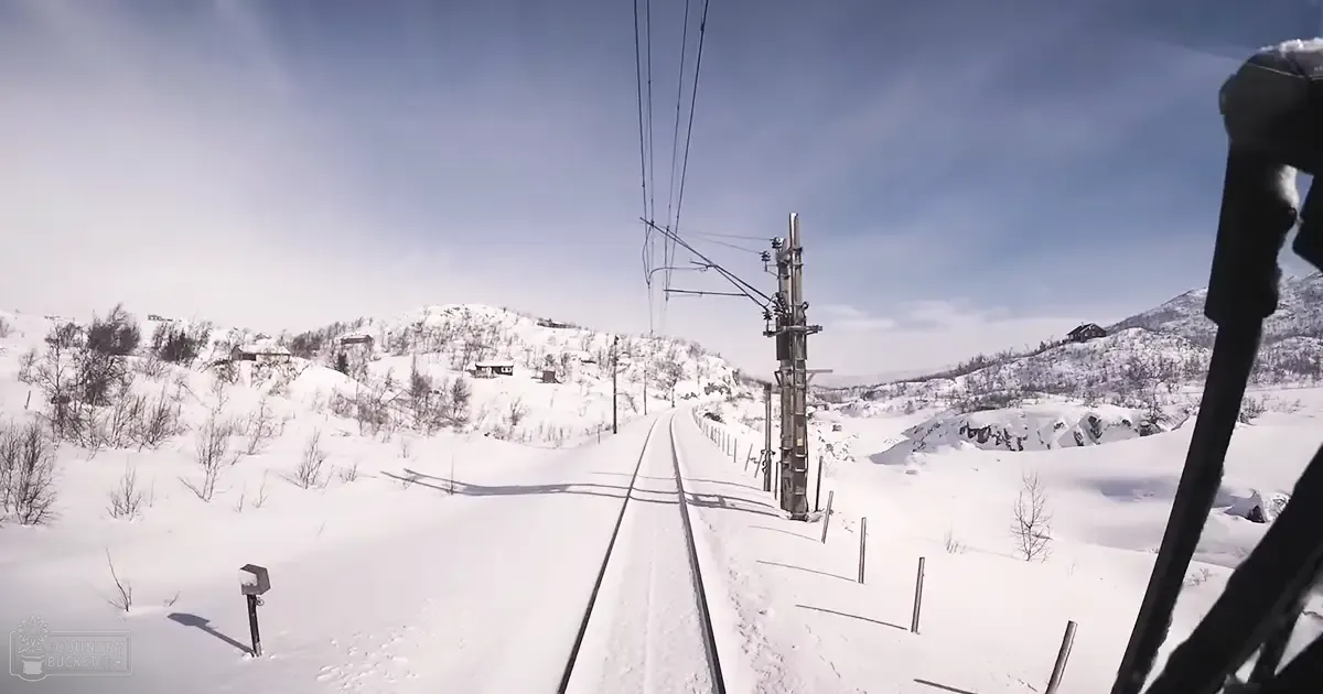 A snowy railway scene in a mountainous area with snow-covered tracks, power lines, distant buildings on hills under a clear blue sky; winter landscape with natural daylight and dominant white, blue, gray, and brown colors.