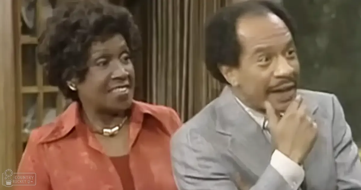 A scene from a 1970s sitcom set in an indoor living room with two characters: a woman wearing a red blouse and necklace, and a man in a light gray suit jacket over a white shirt with cufflinks. A wooden cabinet with glass doors is visible behind the characters.