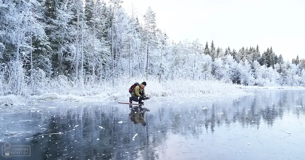 A young adult male in a snowy forest scene ice fishing on a frozen lake at dusk.