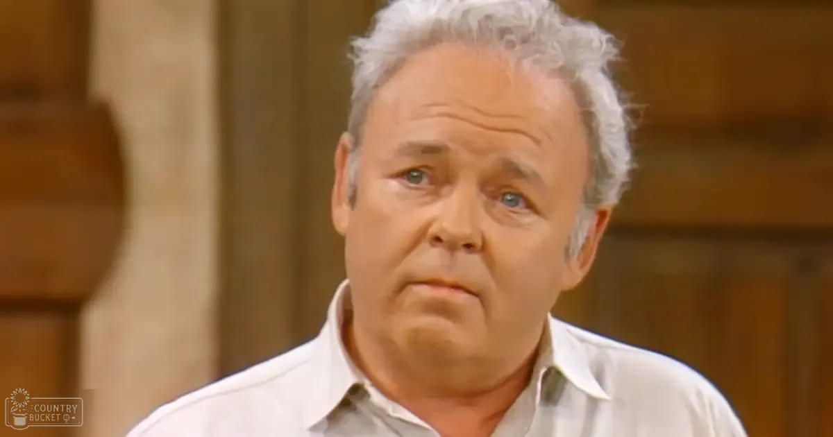 An older adult male wearing a light-colored, collared shirt in an indoor home setting with natural lighting and beige tones.