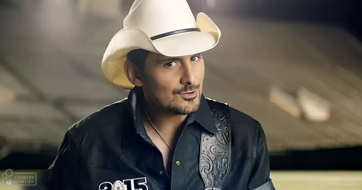 An adult male in a fitted black collared shirt with detailed embroidery, wearing a white cowboy hat and standing indoors with rustic wooden flooring.
